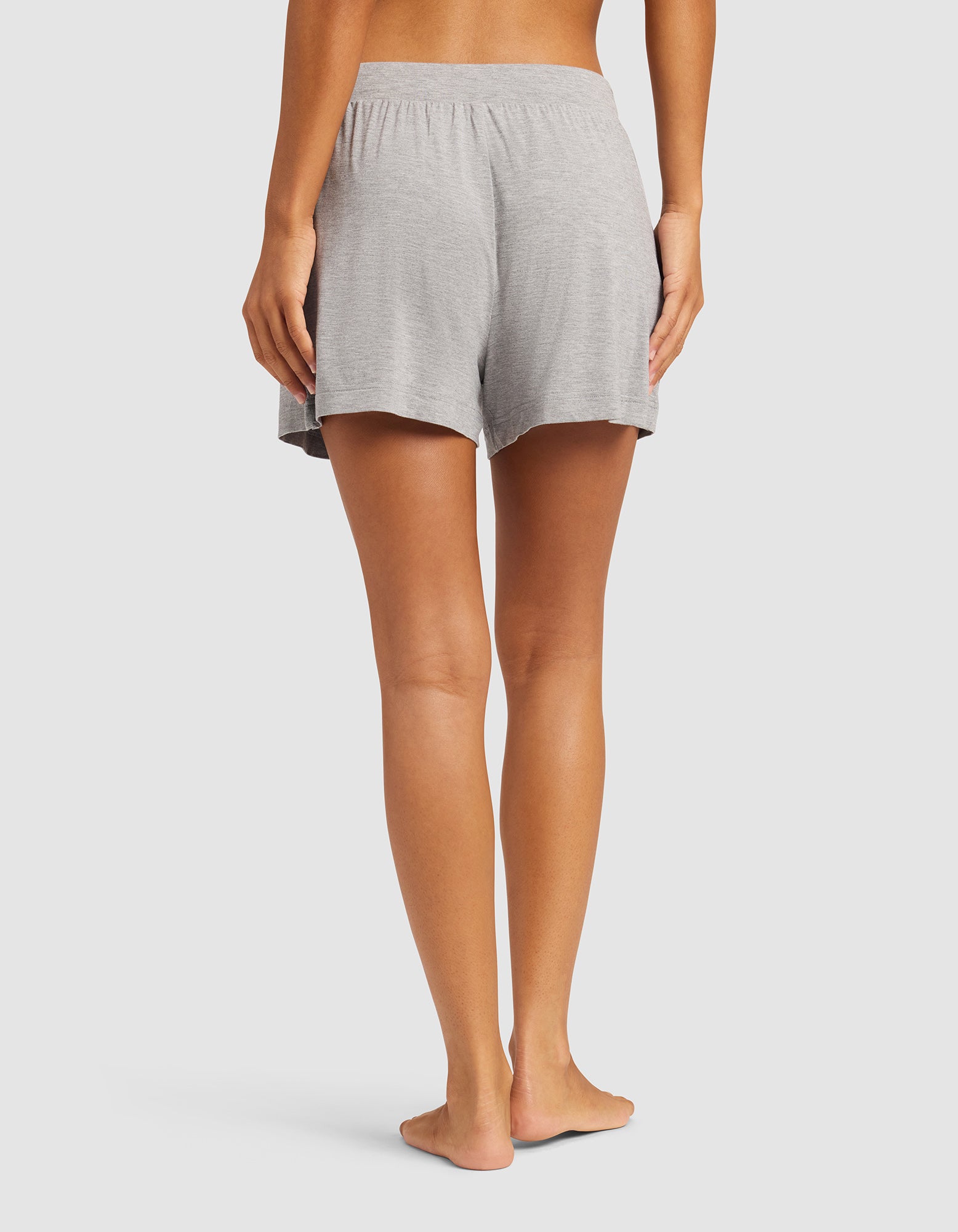 A person stands barefoot on a light background, shown from the waist down and viewed from the back, wearing Cozy Earth Women's Bamboo Stretch-Knit Shorts in light gray.
