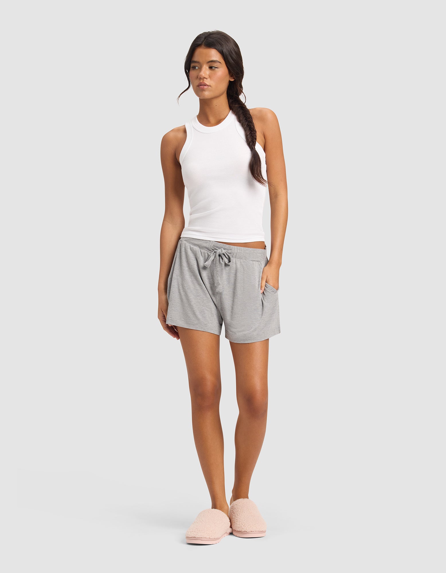 A woman stands against a plain background wearing Cozy Earth's Women's Bamboo Stretch-Knit Shorts in light gray, paired with a white sleeveless tank top and pink fuzzy slippers. Her hair is styled in a loose side braid.