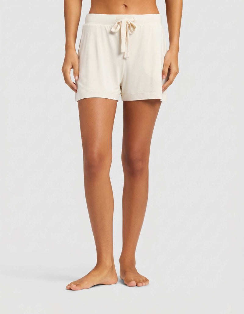 A woman stands barefoot against a plain light background, wearing Cozy Earth's Women's Bamboo Stretch-Knit Shorts in creme. The shorts feature a drawstring waist, relaxed fit, and side pockets.