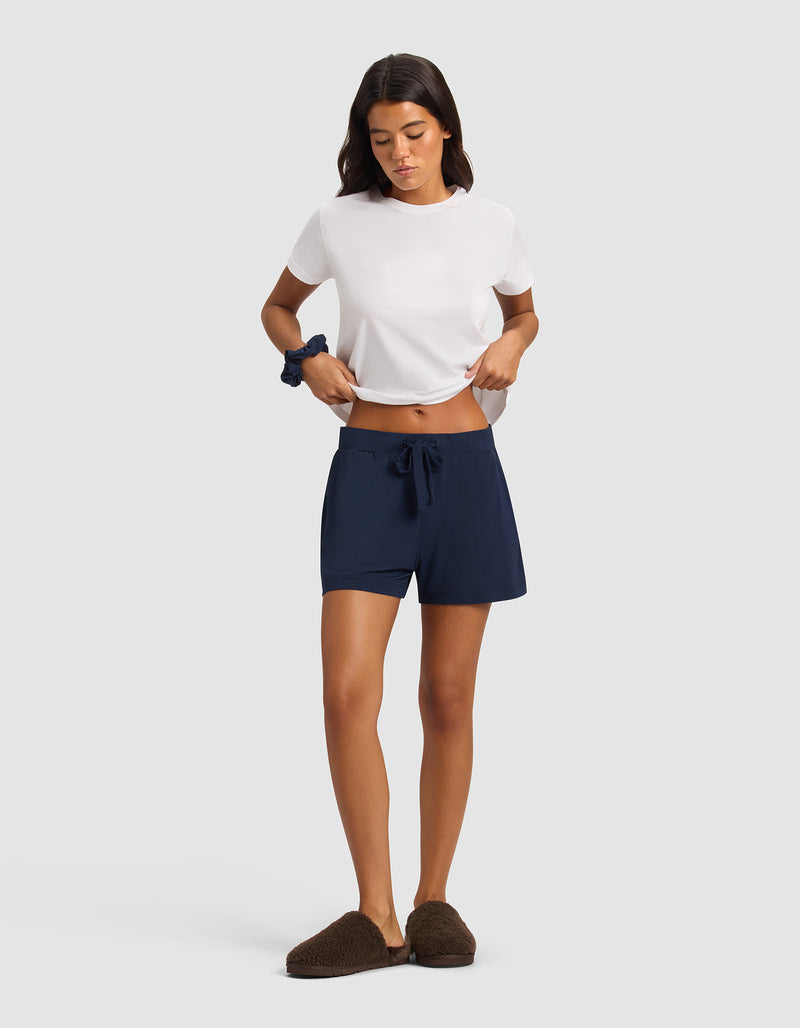 A woman stands against a plain background, wearing Cozy Earth Women's Bamboo Stretch-Knit Shorts in navy blue, a white t-shirt, brown slippers, and a blue scrunchie. She looks down and lightly holds her shirt.