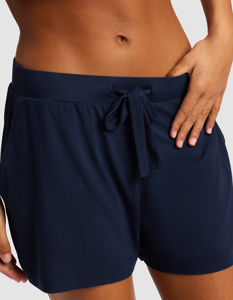 A woman wears Cozy Earth Women's Bamboo Stretch-Knit Shorts in navy blue, featuring a drawstring waistband and side pockets, standing with one hand on her hip against a plain background.