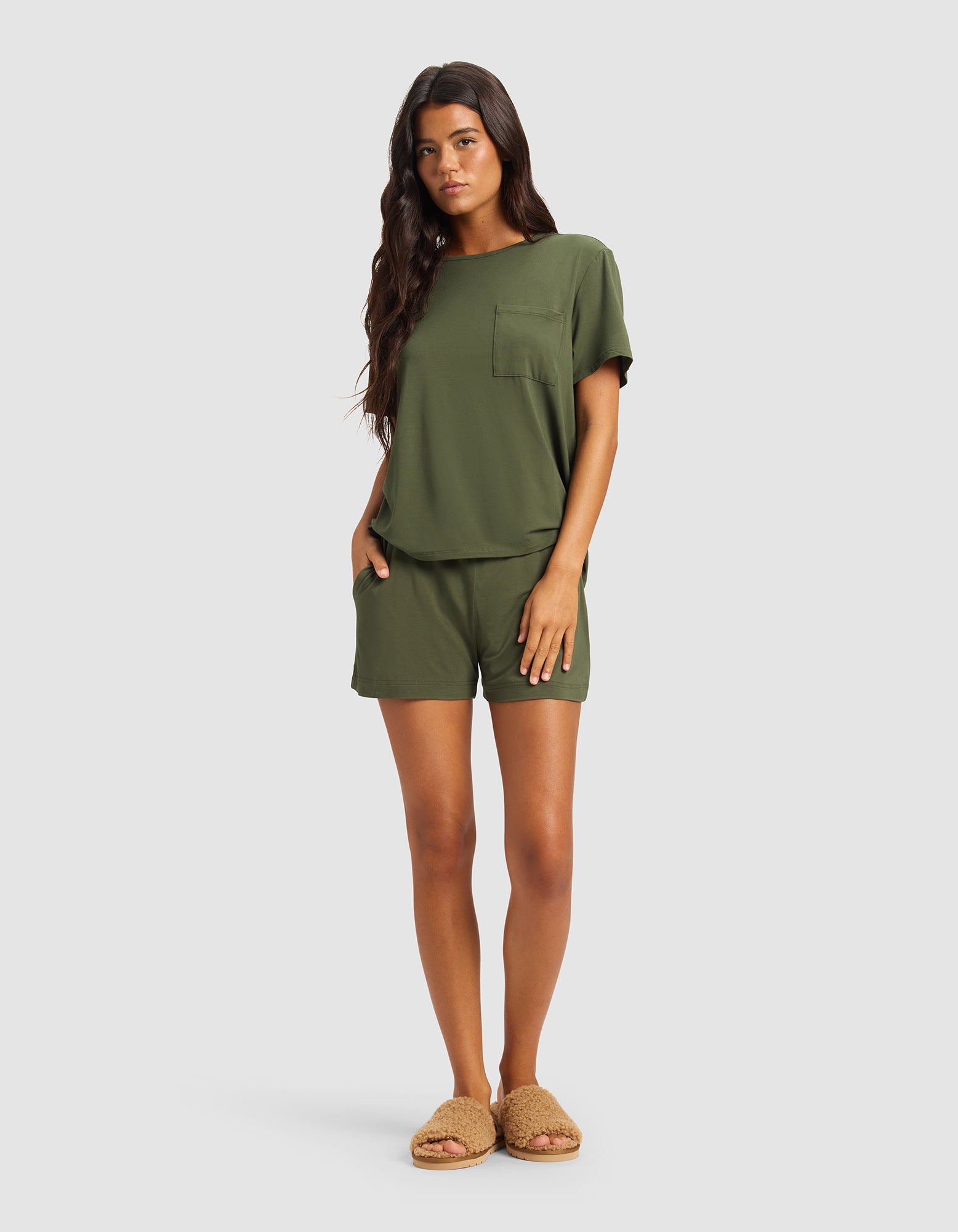 A woman with long dark hair wears Cozy Earth's Women's Bamboo Stretch-Knit Shorts in olive green, paired with a matching T-shirt and tan fuzzy slippers, standing against a light gray background.
