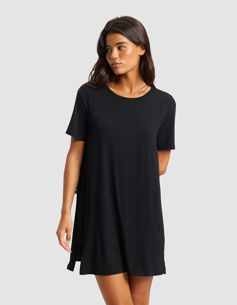 A woman with long brown hair wears the Cozy Earth Women's Bamboo Stretch-Knit Sleep Dress, a short-sleeved, loose-fitting black dress, standing against a plain light gray background and looking slightly to her left.