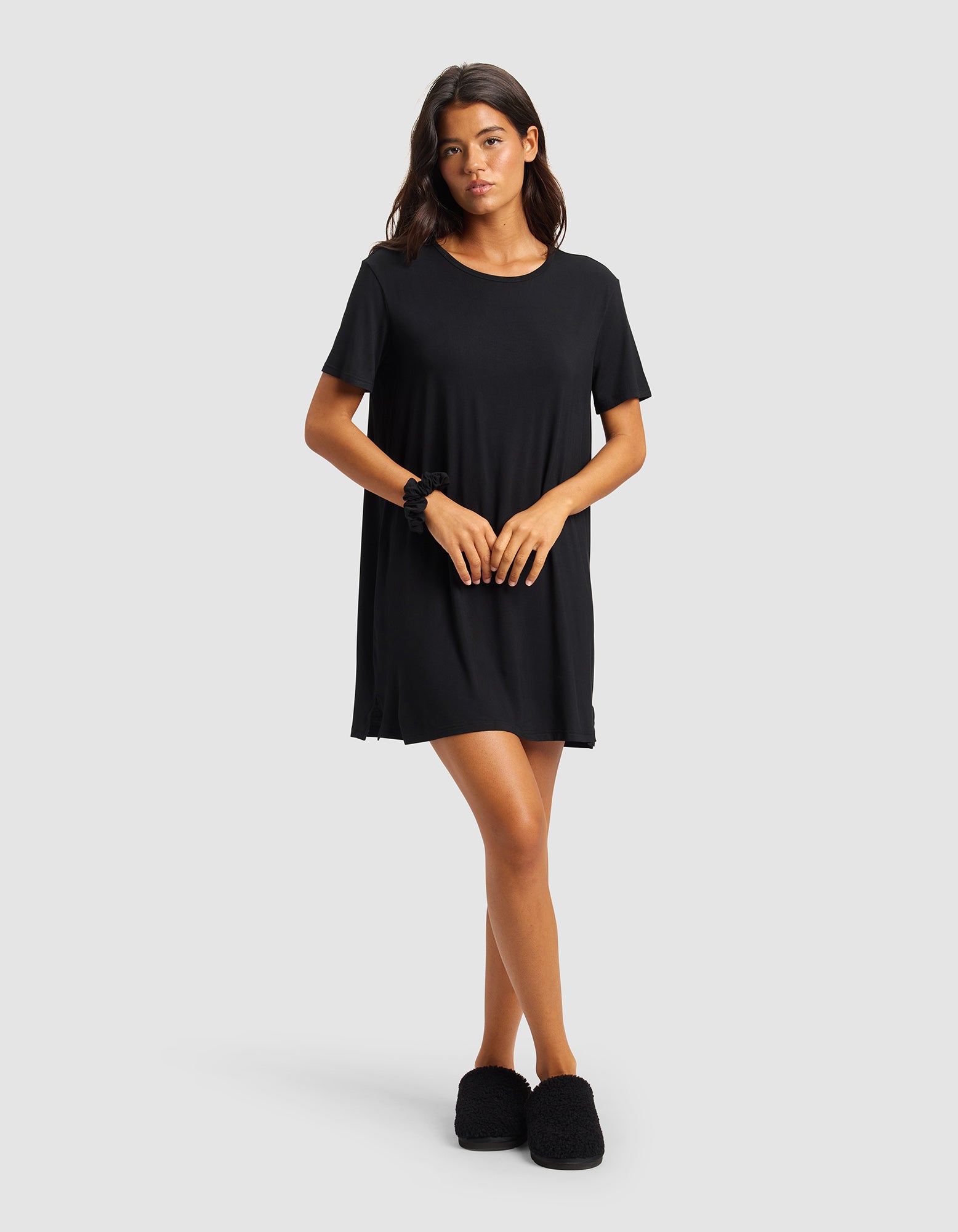 A woman stands against a plain light gray background, wearing Cozy Earth's Women's Bamboo Stretch-Knit Sleep Dress in black and matching slippers. She has long dark hair and her hands are gently clasped in front of her.