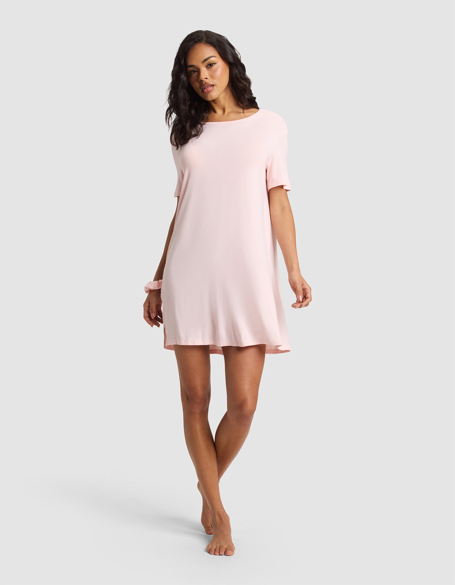 A woman poses barefoot against a light gray background, wearing the Cozy Earth Women's Bamboo Stretch-Knit Sleep Dress in light pink. She has dark wavy hair and a pink scrunchie on her wrist. 
