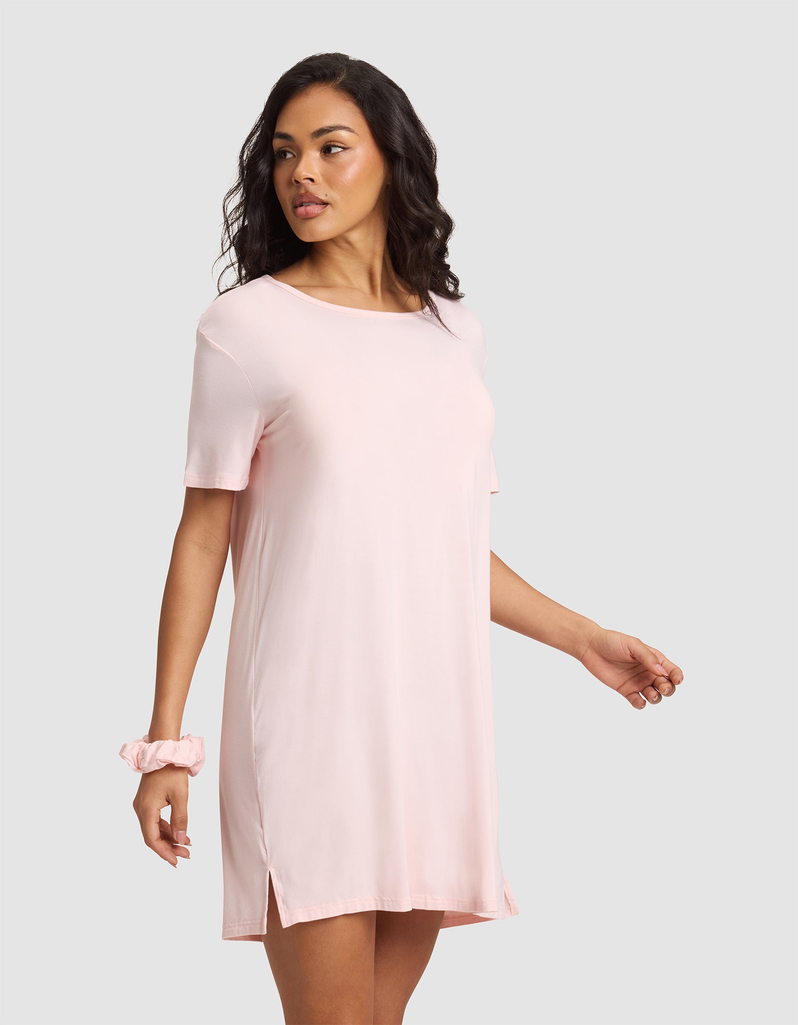 A woman with wavy dark hair and a relaxed expression wears the Cozy Earth Women's Bamboo Stretch-Knit Sleep Dress in light pink. She stands against a plain gray background, with a matching pink scrunchie on her left wrist. |Color:Blossom