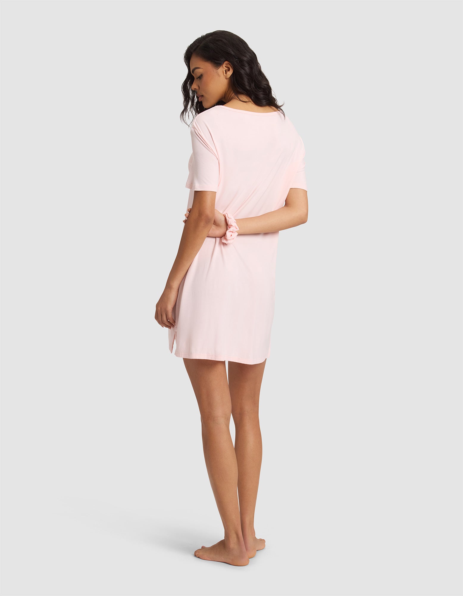 A woman with long dark hair stands barefoot, facing away in a light pink Cozy Earth Women's Bamboo Stretch-Knit Sleep Dress, with one hand behind her back against a plain grey background. 