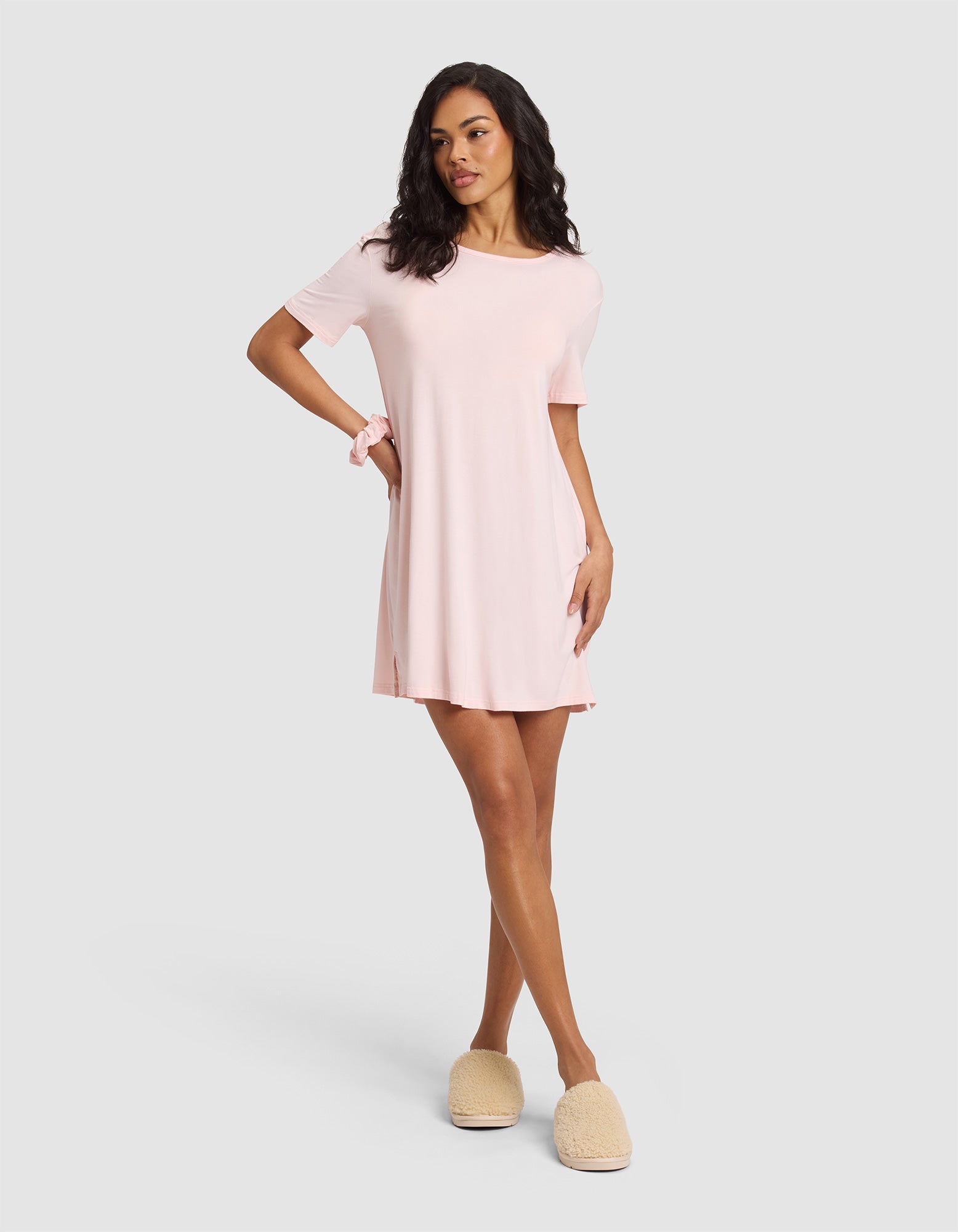 A woman poses in a relaxed stance wearing the Cozy Earth Women's Bamboo Stretch-Knit Sleep Dress in light pink and beige fuzzy slippers, with one hand on her hip against a plain light gray background. 