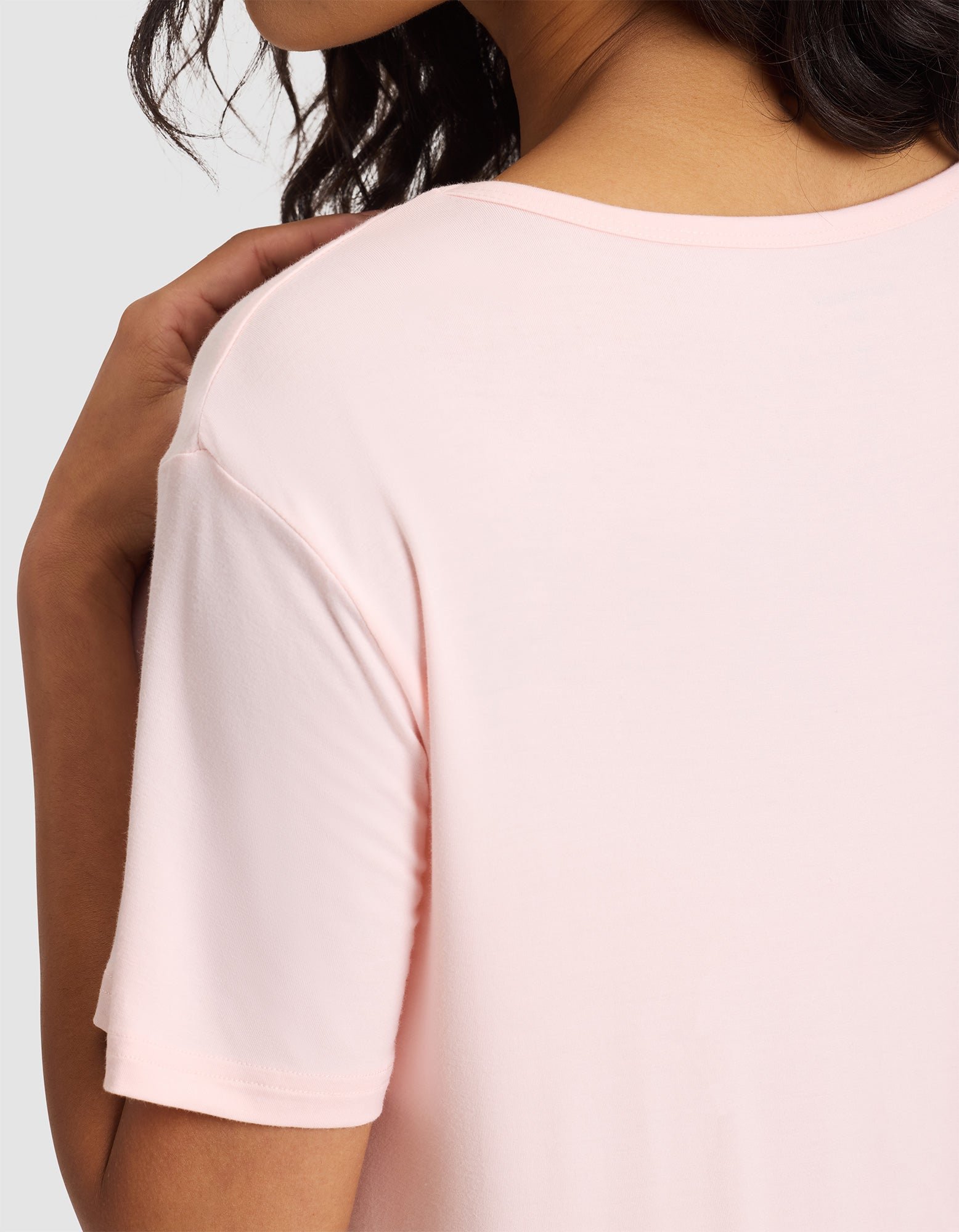 A woman with wavy dark hair models the Cozy Earth Women's Bamboo Stretch-Knit Sleep Dress in light pink, highlighting her shoulder and upper arm against a plain gray background. 