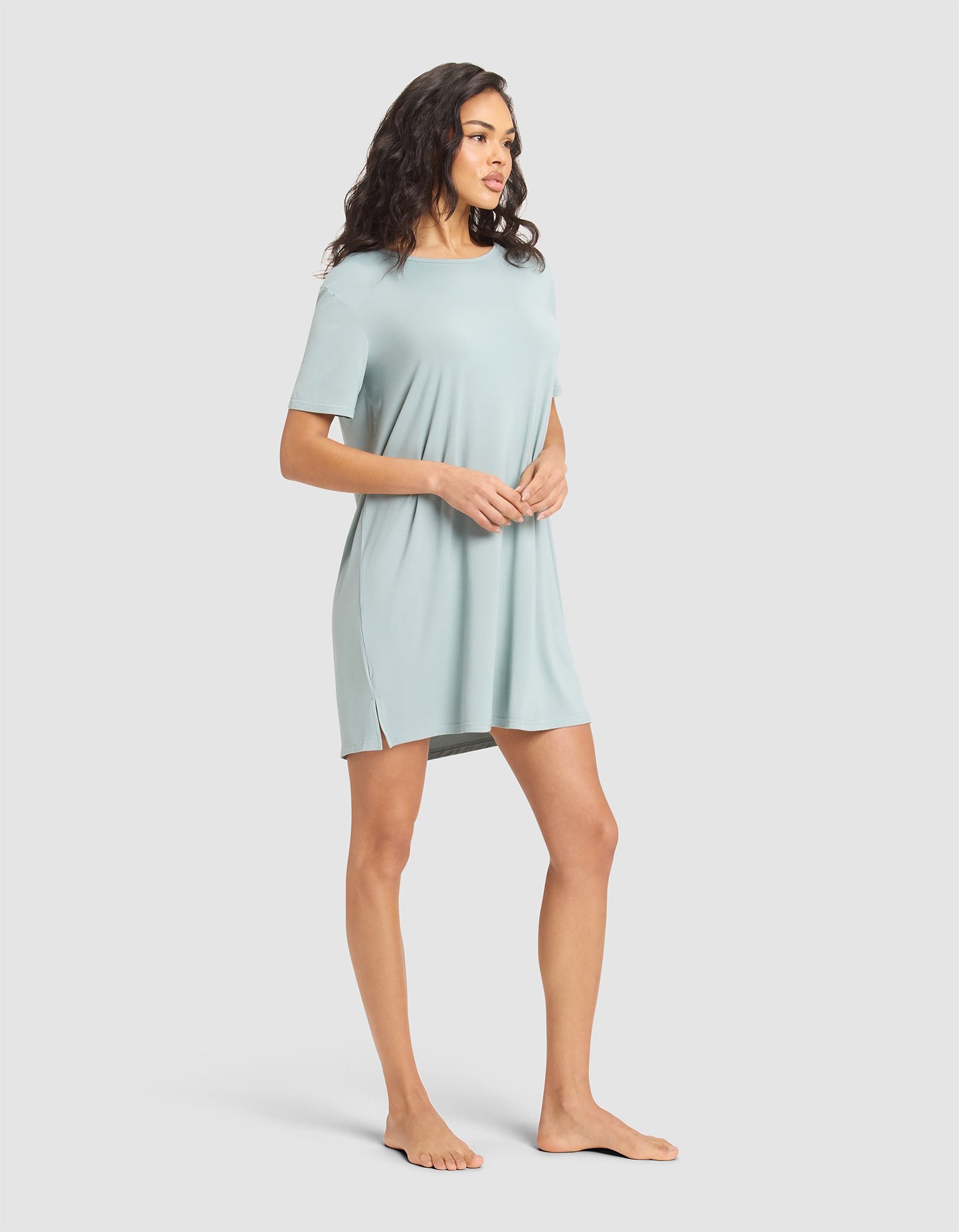 A woman stands barefoot against a plain background, wearing the Cozy Earth Women's Bamboo Stretch-Knit Sleep Dress—a relaxed-fit, short-sleeved, light blue dress with side slits—looking to her left with hands clasped in front. 