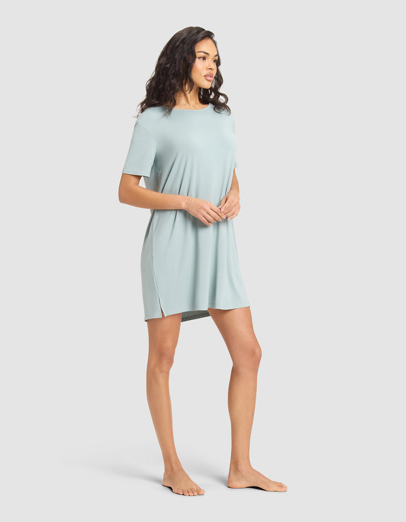 A woman stands barefoot against a plain background, wearing the Cozy Earth Women's Bamboo Stretch-Knit Sleep Dress—a relaxed-fit, short-sleeved, light blue dress with side slits—looking to her left with hands clasped in front.