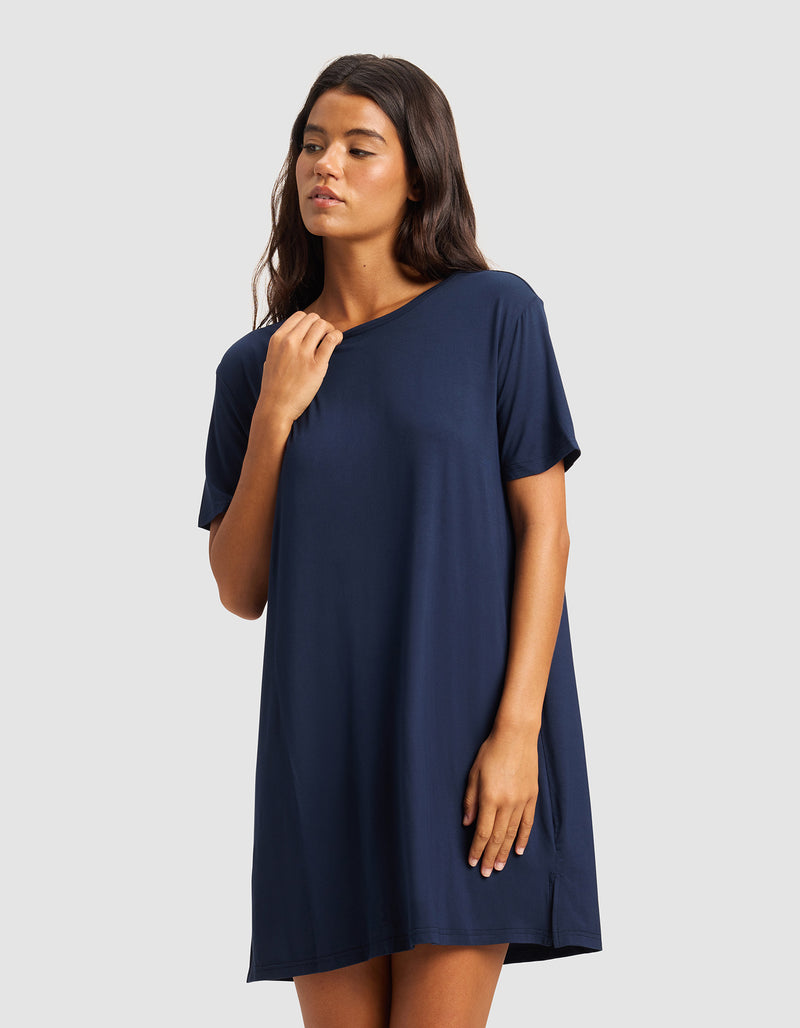 A woman stands against a light gray background, wearing Cozy Earth's Women's Bamboo Stretch-Knit Sleep Dress in navy blue. With long dark hair, she gazes to the side and lightly touches her collar.