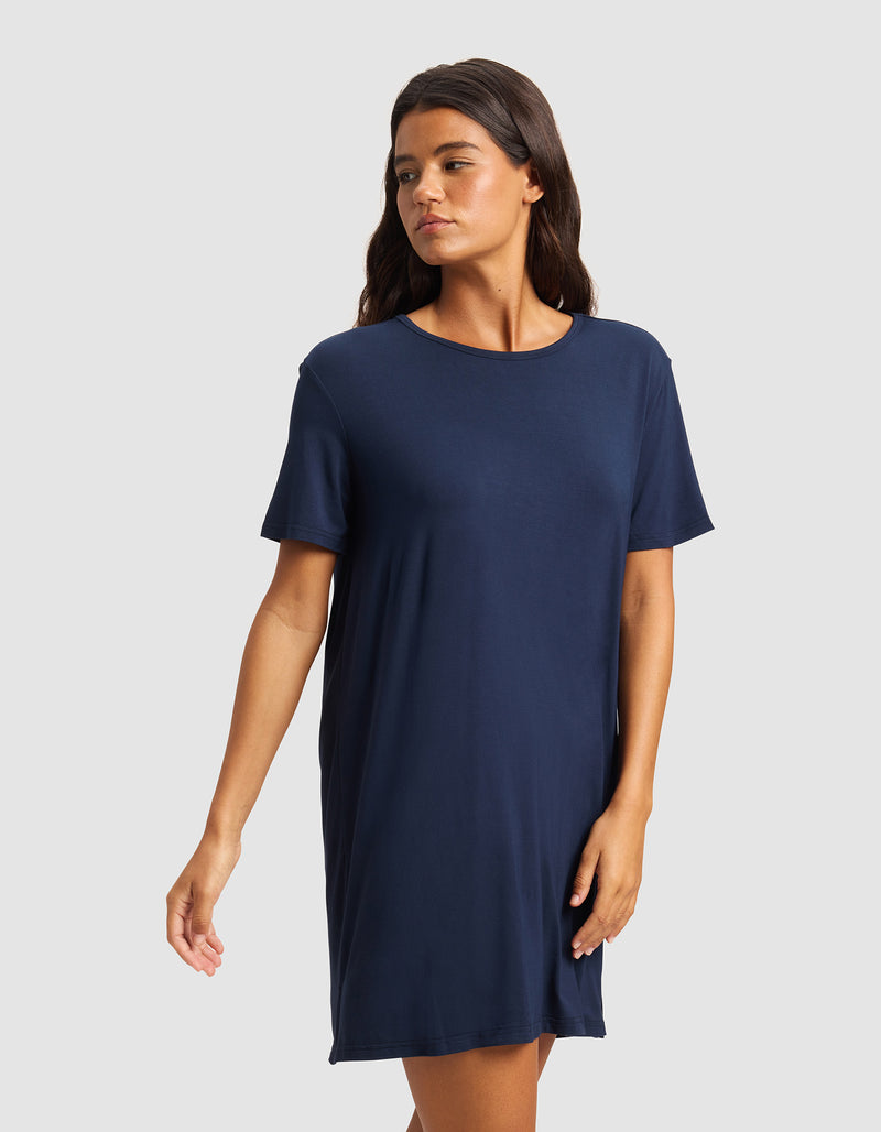 A woman models the Cozy Earth Women's Bamboo Stretch-Knit Sleep Dress, a loose-fitting, short-sleeve navy blue piece, while standing against a light gray background and gazing to the side.