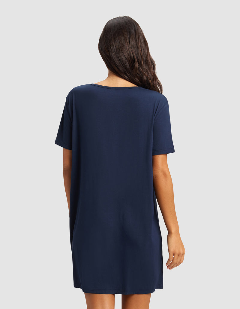 A woman with long wavy hair stands facing away, wearing the Cozy Earth Women's Bamboo Stretch-Knit Sleep Dress in navy blue against a plain light gray background.