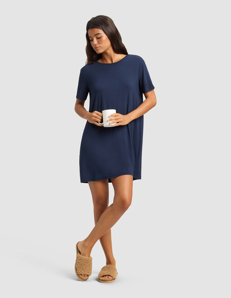 A woman in a Cozy Earth Women's Bamboo Stretch-Knit Sleep Dress stands holding a white mug, wearing tan fuzzy slippers. She has long dark hair and looks relaxed against a plain light gray background.