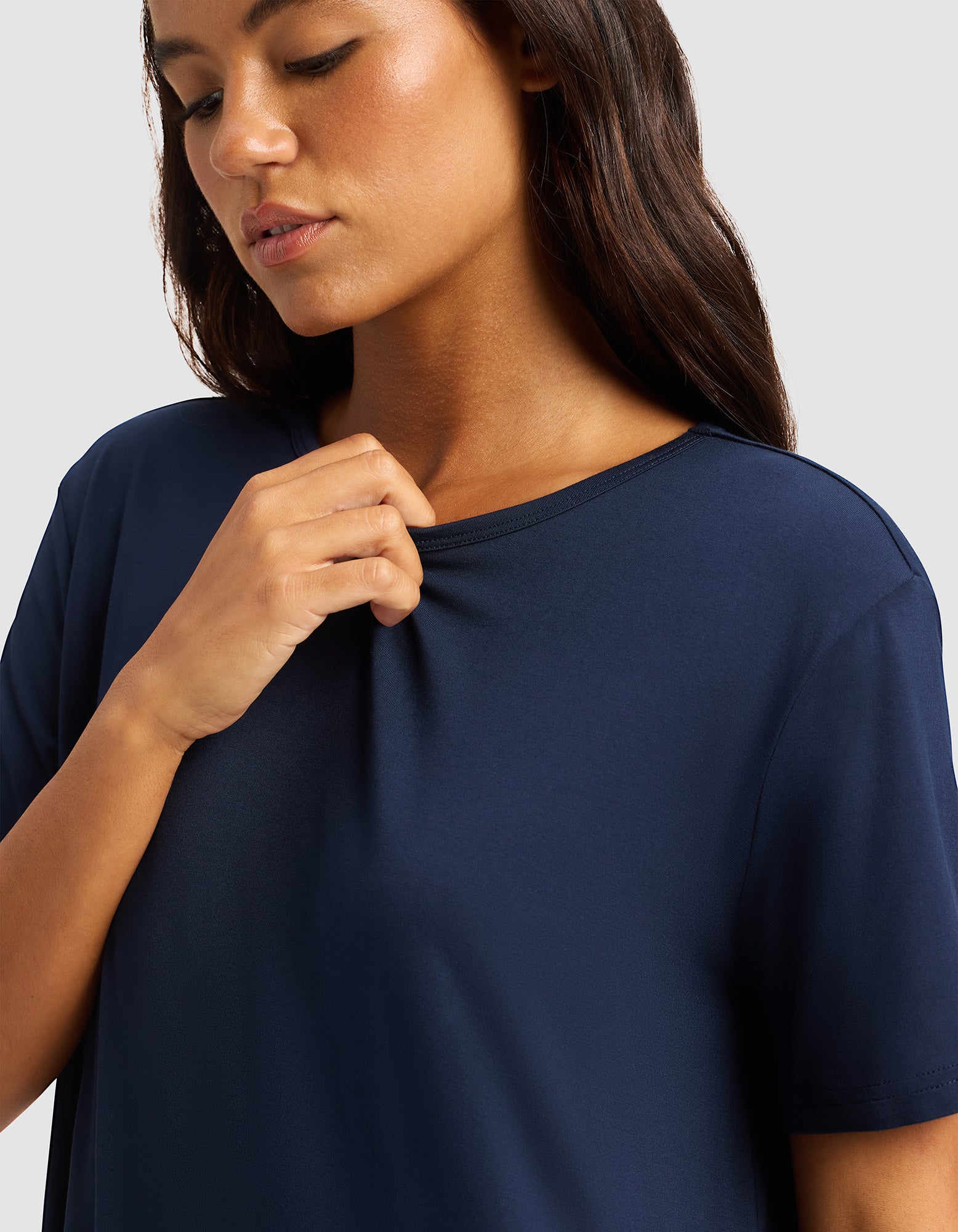 A woman with long brown hair wears the Cozy Earth Women's Bamboo Stretch-Knit Sleep Dress in navy blue, gently holding the collar with her right hand and looking downward calmly against a plain light gray background.