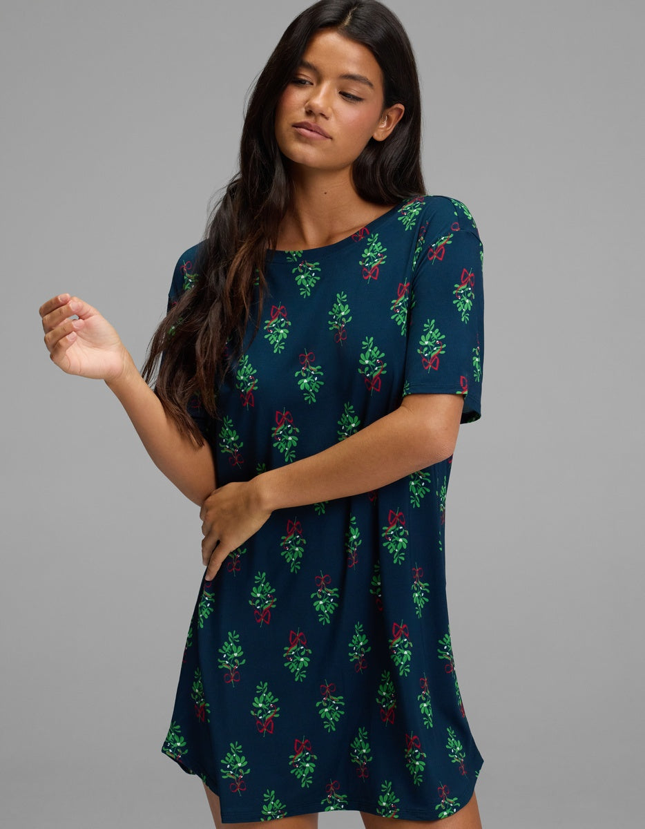A woman wears a dark teal Cozy Earth Women’s Bamboo Stretch-Knit Sleep Dress featuring a repeating pattern of green mistletoe sprigs and red bows.