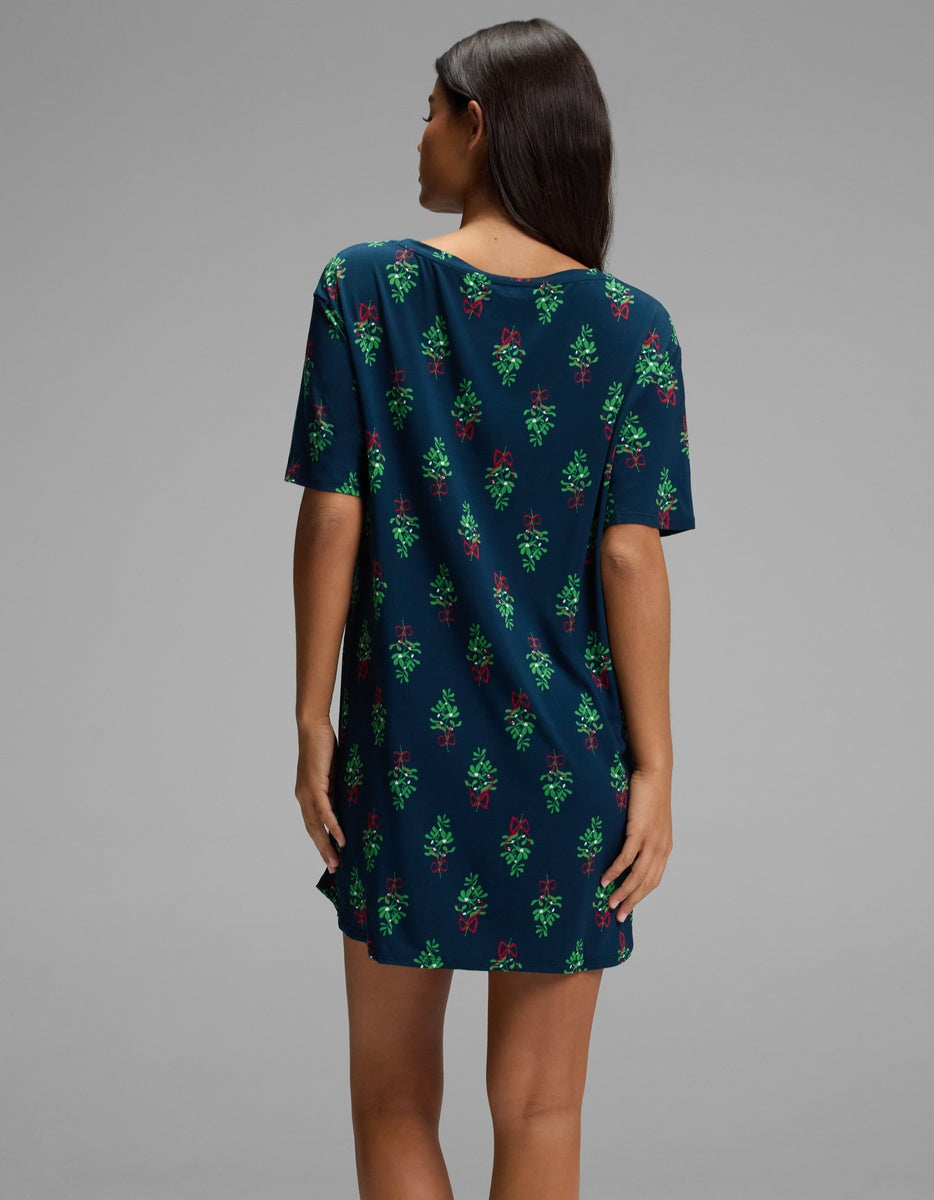 Back view of a person wearing a dark blue Cozy Earth Women’s Bamboo Stretch-Knit Sleep Dress featuring a short-sleeved silhouette with a pattern of mistletoe sprigs and red bows.