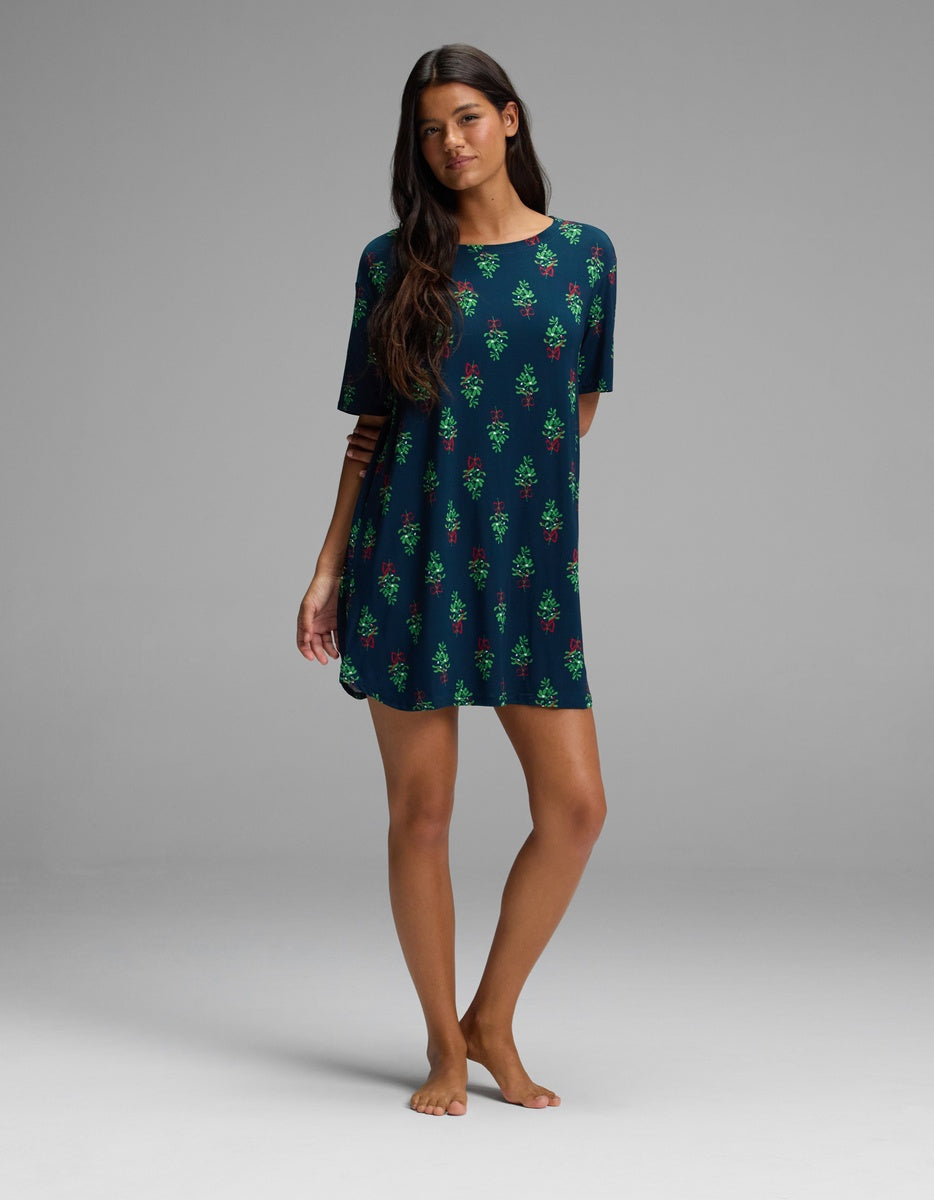 A woman wears a dark blue Cozy Earth Women’s Bamboo Stretch-Knit Sleep Dress featuring a short-sleeved design with a repeating pattern of green Christmas trees and red bows.