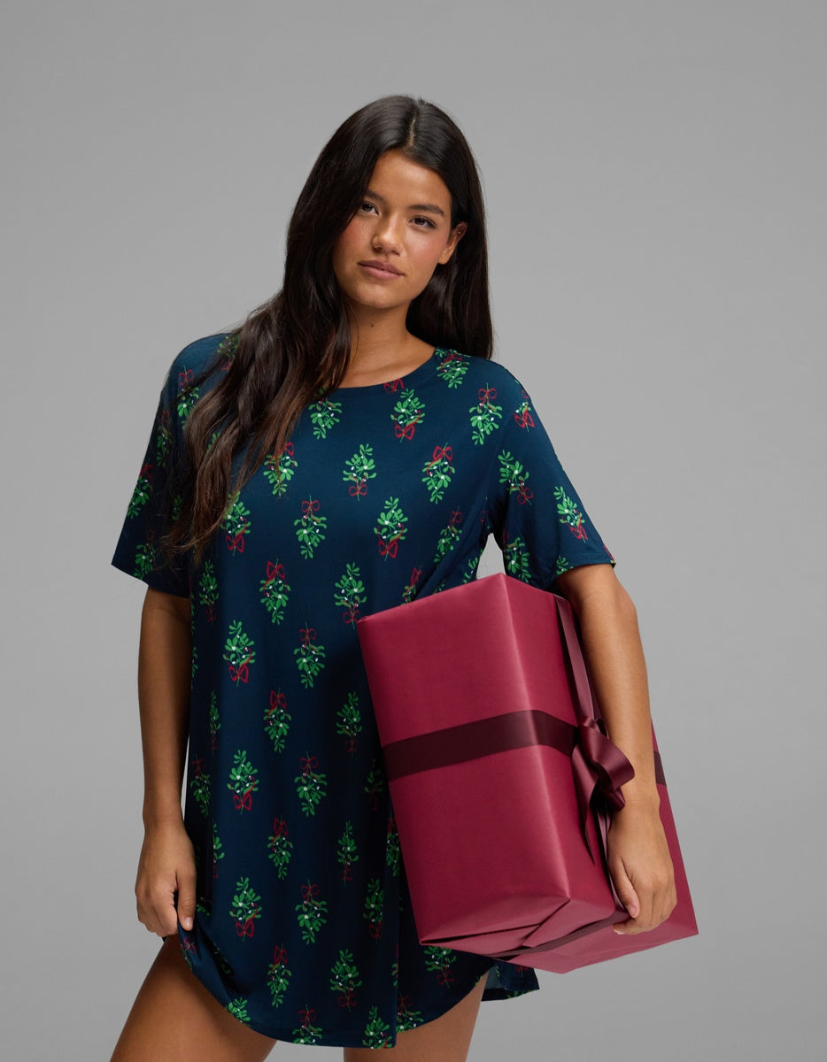 A woman wearing a navy blue Women’s Bamboo Stretch-Knit Sleep Dress with a green mistletoe pattern holds a large gift wrapped in burgundy paper.
