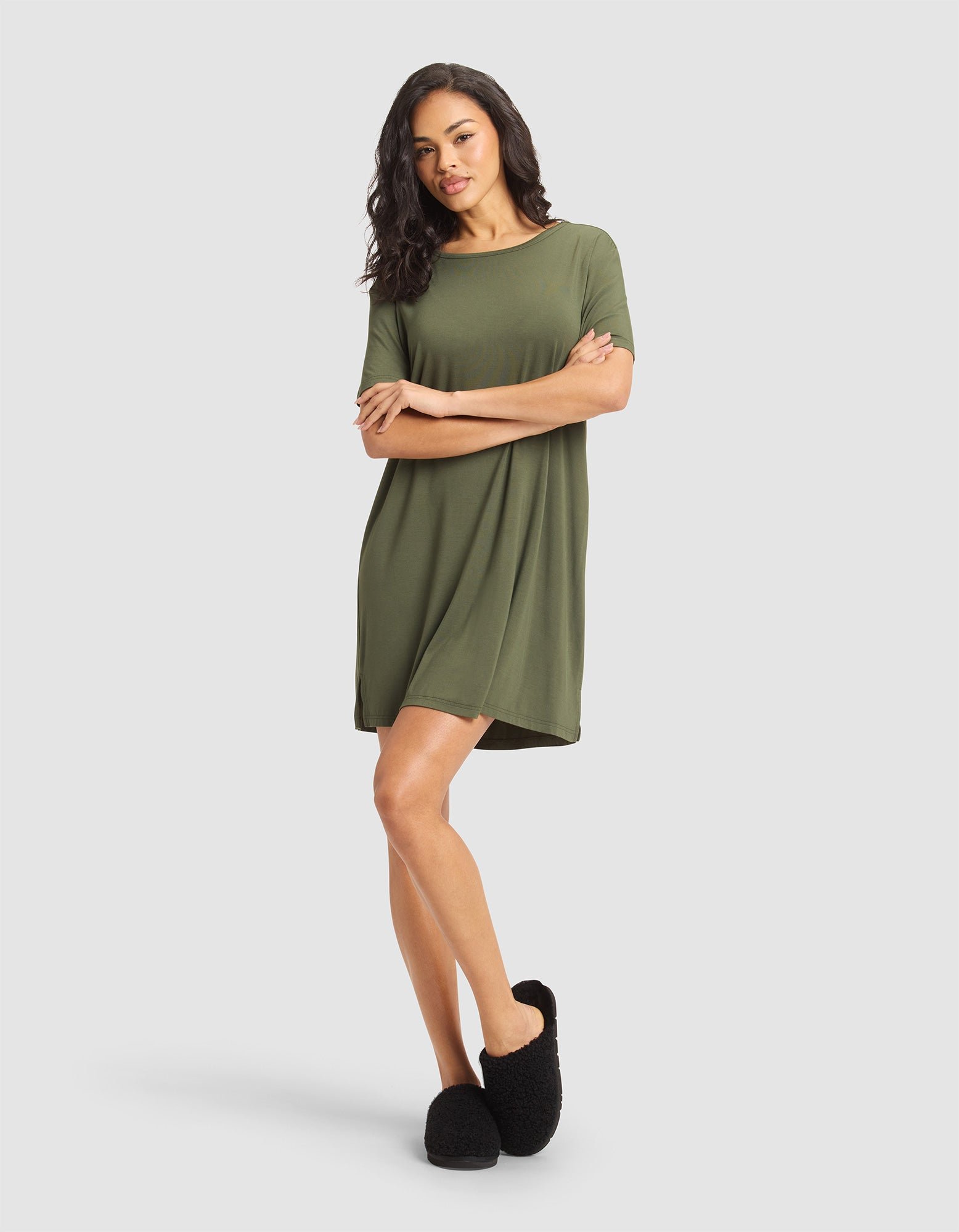 A woman with long, wavy hair wears the Cozy Earth Women's Bamboo Stretch-Knit Sleep Dress in olive green and black fuzzy slippers, standing against a plain light gray background with one leg slightly crossed. 
