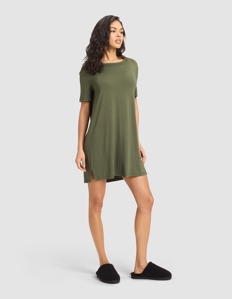 A woman poses against a plain background wearing the Cozy Earth Women's Bamboo Stretch-Knit Sleep Dress in olive green, featuring short sleeves and side slits. She stands slightly sideways with her left leg forward and right arm relaxed.
