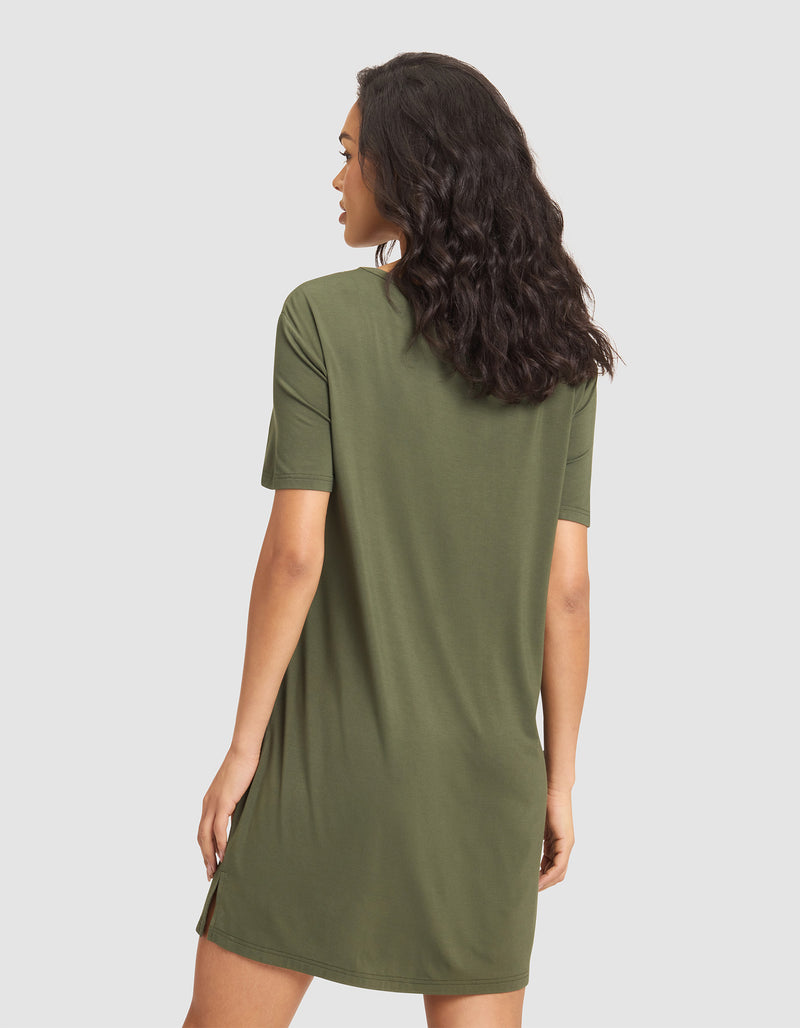 A woman with wavy dark hair faces away, wearing the Cozy Earth Women's Bamboo Stretch-Knit Sleep Dress in olive green. The short-sleeve, knee-length dress stands out against a plain light gray background.