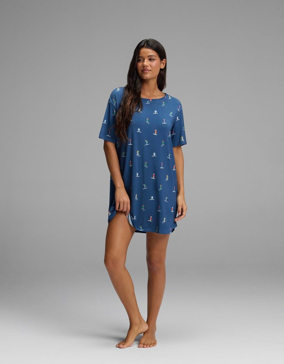 A woman wears a blue short-sleeve Cozy Earth Women’s Bamboo Stretch-Knit Sleep Dress patterned with small skiers.