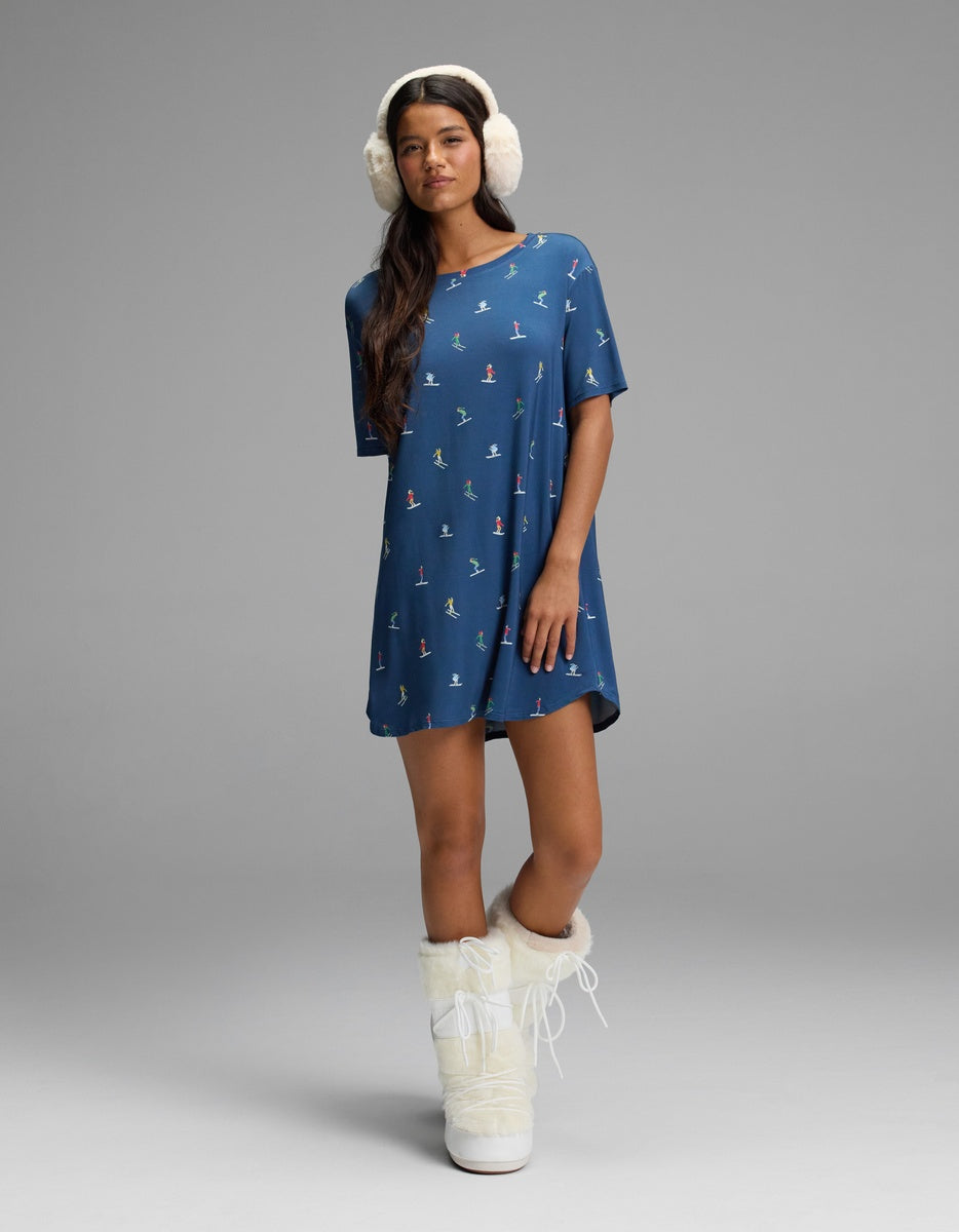 A woman wears a blue short-sleeved Cozy Earth Women’s Bamboo Stretch-Knit Sleep Dress with a small winter sports print, white earmuffs, and white faux-fur snow boots.
