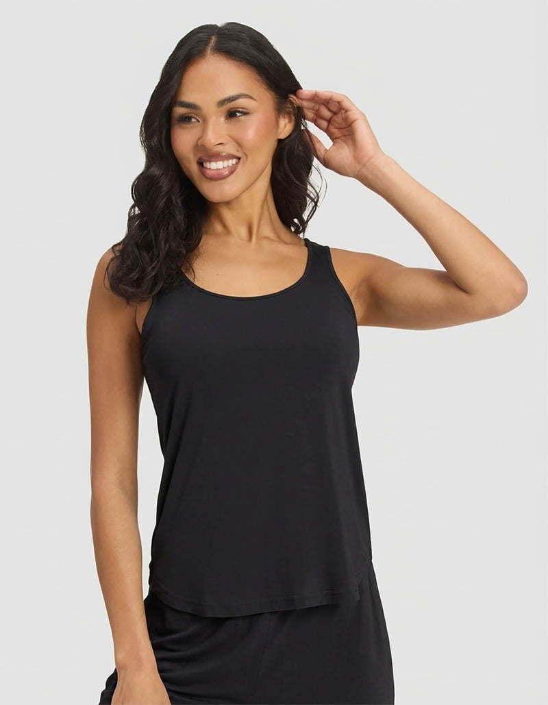 A woman with long, wavy dark hair smiles and poses in Cozy Earth's Women's Bamboo Stretch-Knit Tank & Short Set, featuring a sleeveless black tank top and matching shorts, against a plain light background.