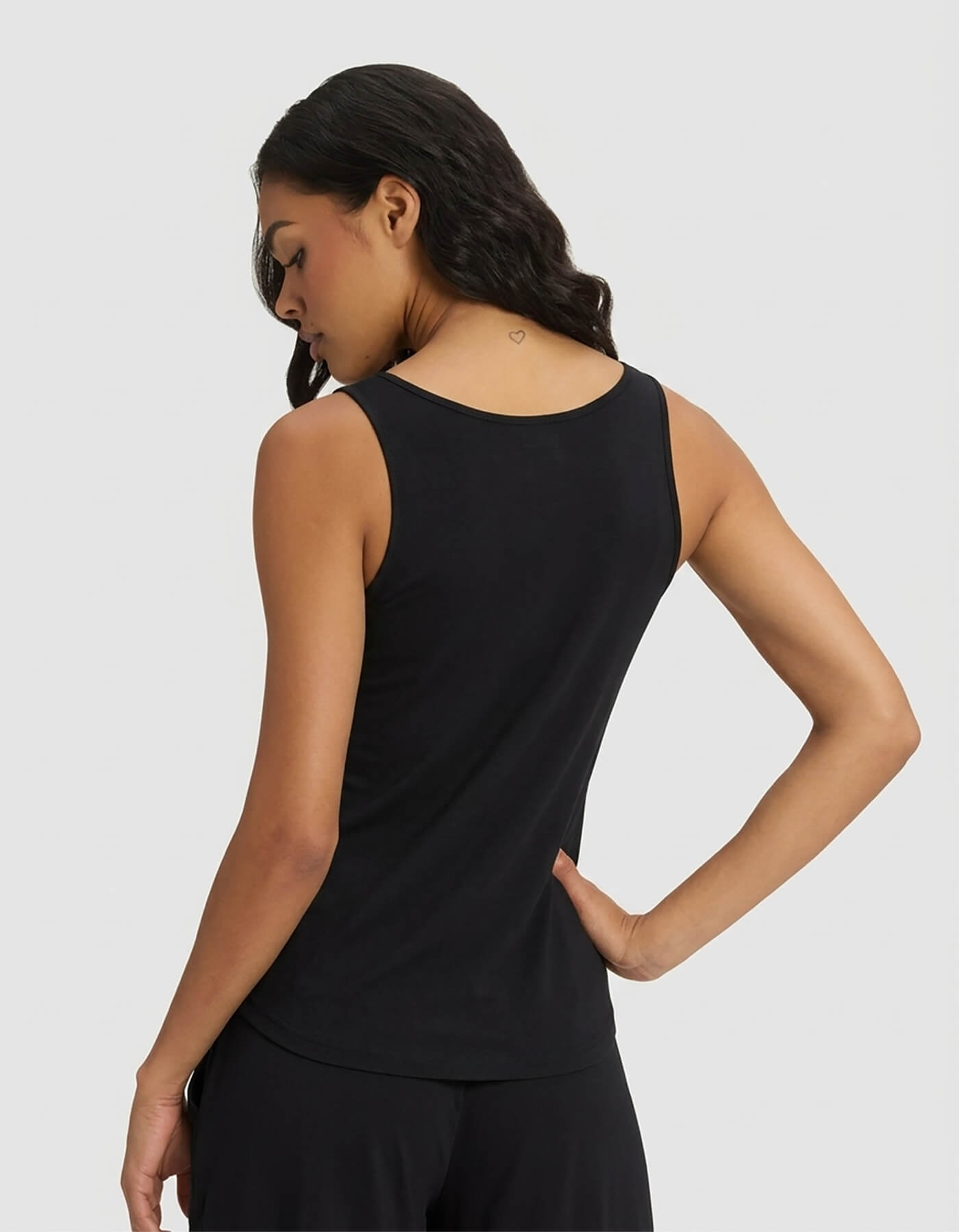 A woman with wavy dark hair models the Cozy Earth Women’s Bamboo Stretch-Knit Tank & Short Set in black, standing against a light gray background. She faces away, looking down, revealing a heart tattoo on the back of her neck.