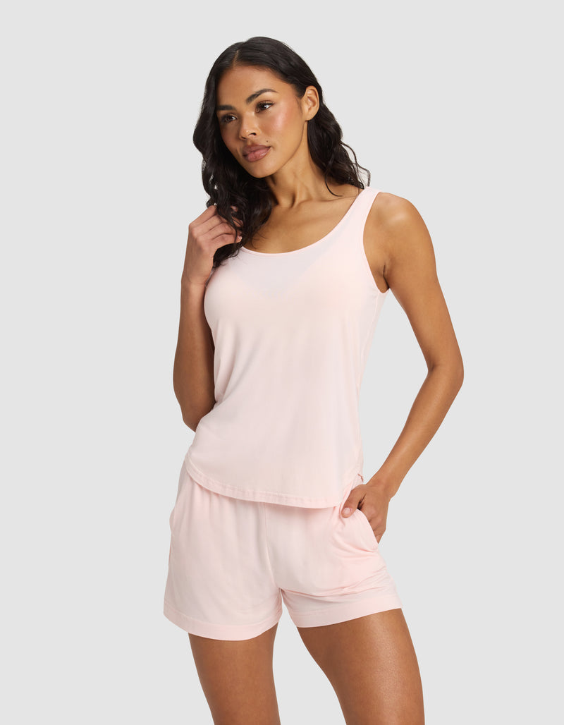 A woman with long wavy dark hair stands against a light gray background, wearing the Cozy Earth Women's Bamboo Stretch-Knit Lounge Tank in light pink and matching shorts, with one hand in her pocket, looking slightly to the side.