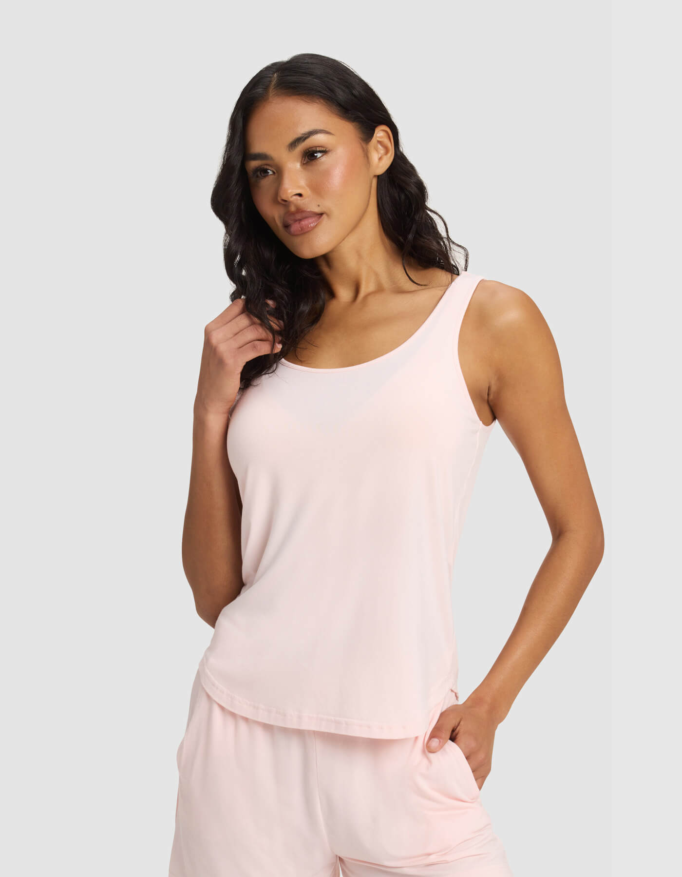 A woman with long, wavy dark hair wears the Cozy Earth Women's Bamboo Stretch-Knit Tank & Tapered Pant Set in light pink, standing against a plain light gray background with one hand in her pocket.