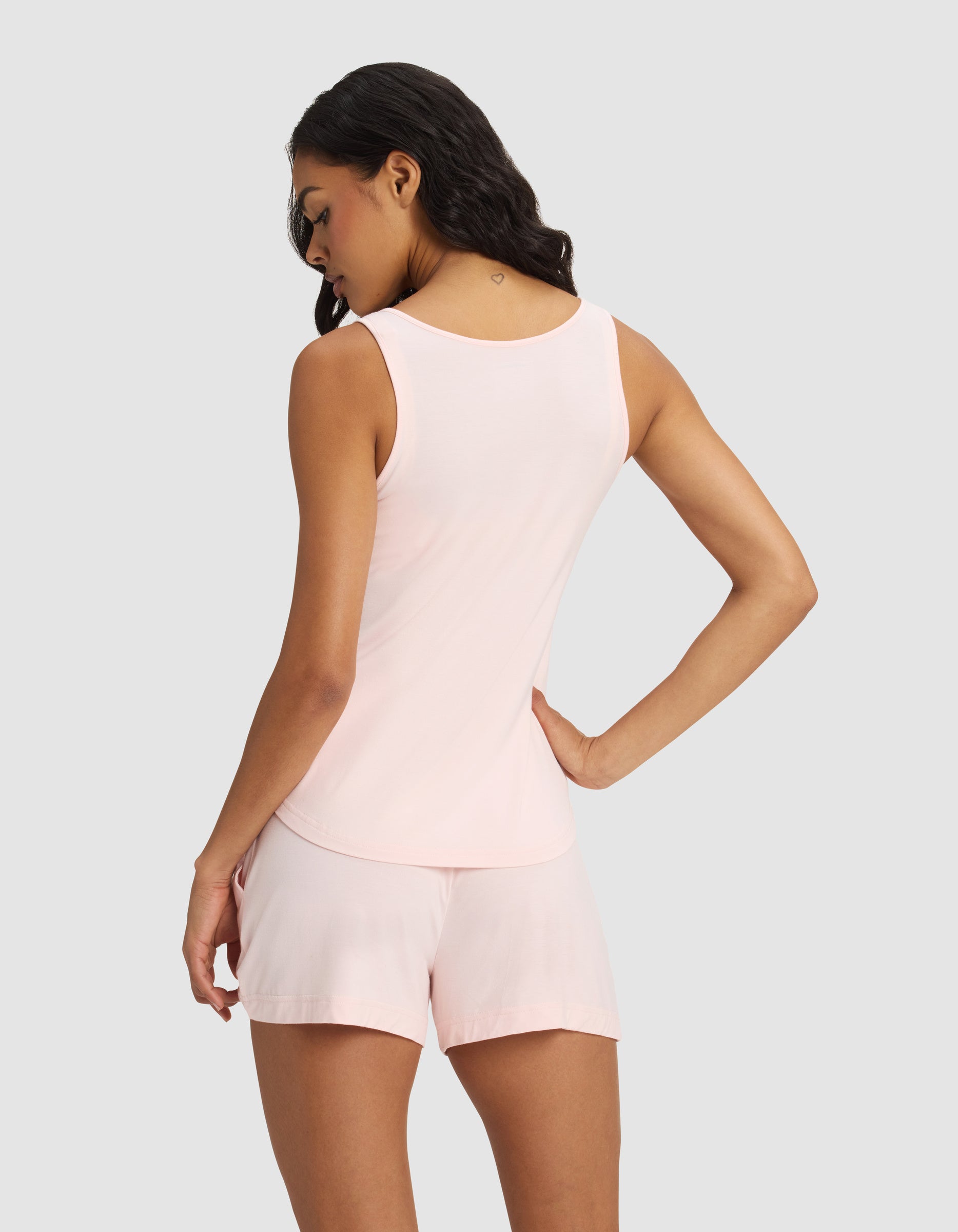 A woman stands with her back to the camera, wearing the Cozy Earth Women's Bamboo Stretch-Knit Lounge Tank in light pink, paired with matching shorts, against a plain light gray background.|Color:Blossom