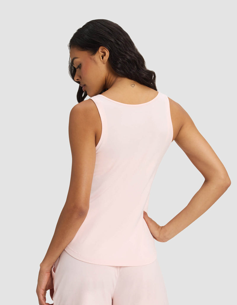A woman with long dark hair faces away, showcasing a small heart tattoo and wearing Cozy Earth's Women's Bamboo Stretch-Knit Tank & Tapered Pant Set in light pink. The background is light gray.