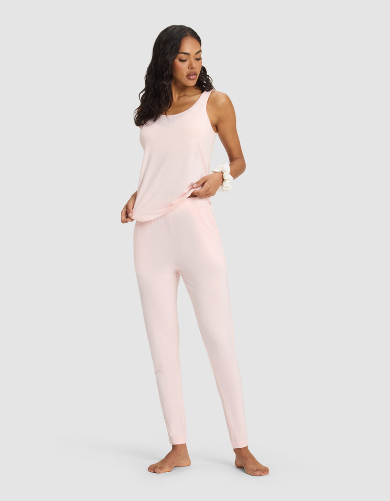 A woman stands barefoot against a plain background, wearing the Cozy Earth Women's Bamboo Stretch-Knit Lounge Tank in light pink with fitted pants. She has long, wavy hair and holds the hem of her top with one hand.