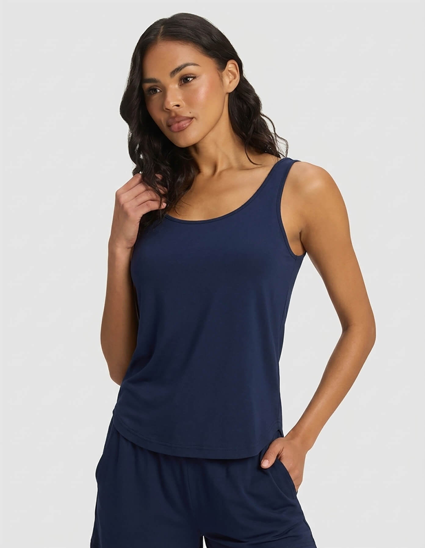 A woman wears the Cozy Earth Women's Bamboo Stretch-Knit Tank & Short Set in navy blue, standing against a light gray background with one hand in her pocket and the other touching her hair, smiling softly.