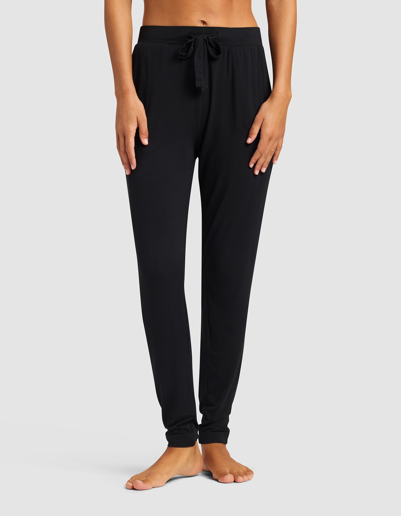 A woman stands barefoot against a light gray background, wearing Cozy Earth's Women's Bamboo Stretch-Knit Tapered Pant in black. Her hands rest at her sides, and the focus is on the pants from waist to ankles.