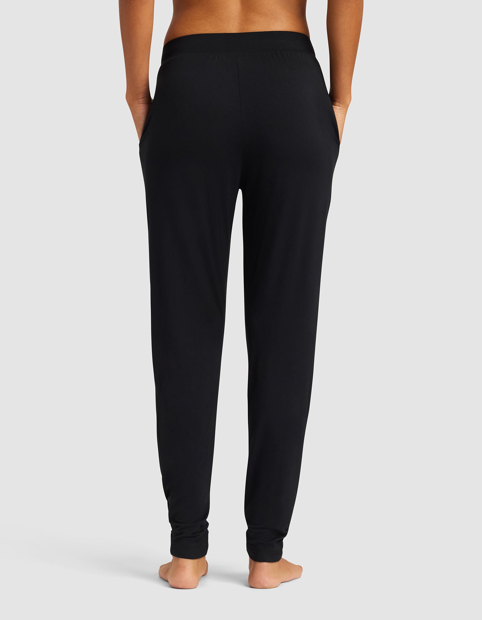 A woman stands barefoot facing away from the camera, wearing Cozy Earth's Women's Bamboo Stretch-Knit Tapered Pant in black with hands in pockets against a plain light gray background.