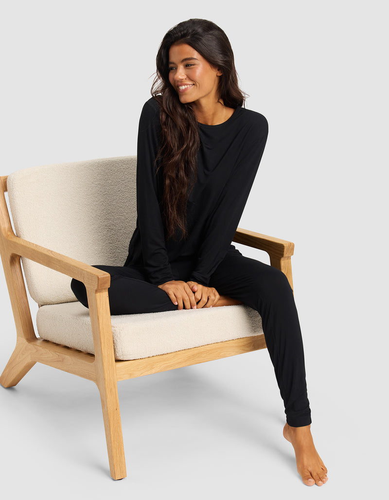 A woman with long dark hair sits barefoot on a light wooden chair, smiling and looking to the side while wearing Cozy Earth Women's Bamboo Stretch-Knit Tapered Pants and a matching black long-sleeve top.