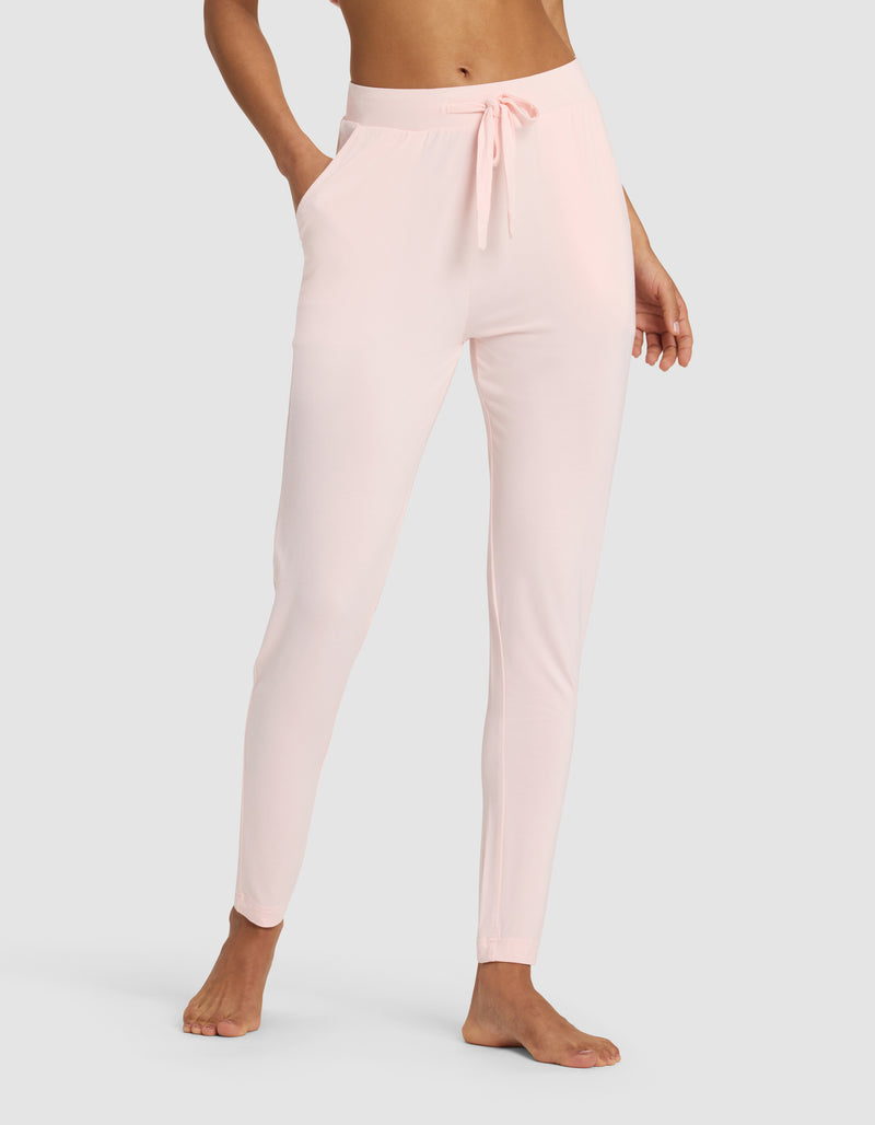 A woman wears Cozy Earth's Women's Bamboo Stretch-Knit Tapered Pant in light pink, featuring a drawstring waist and side pockets, standing barefoot against a plain light gray background.