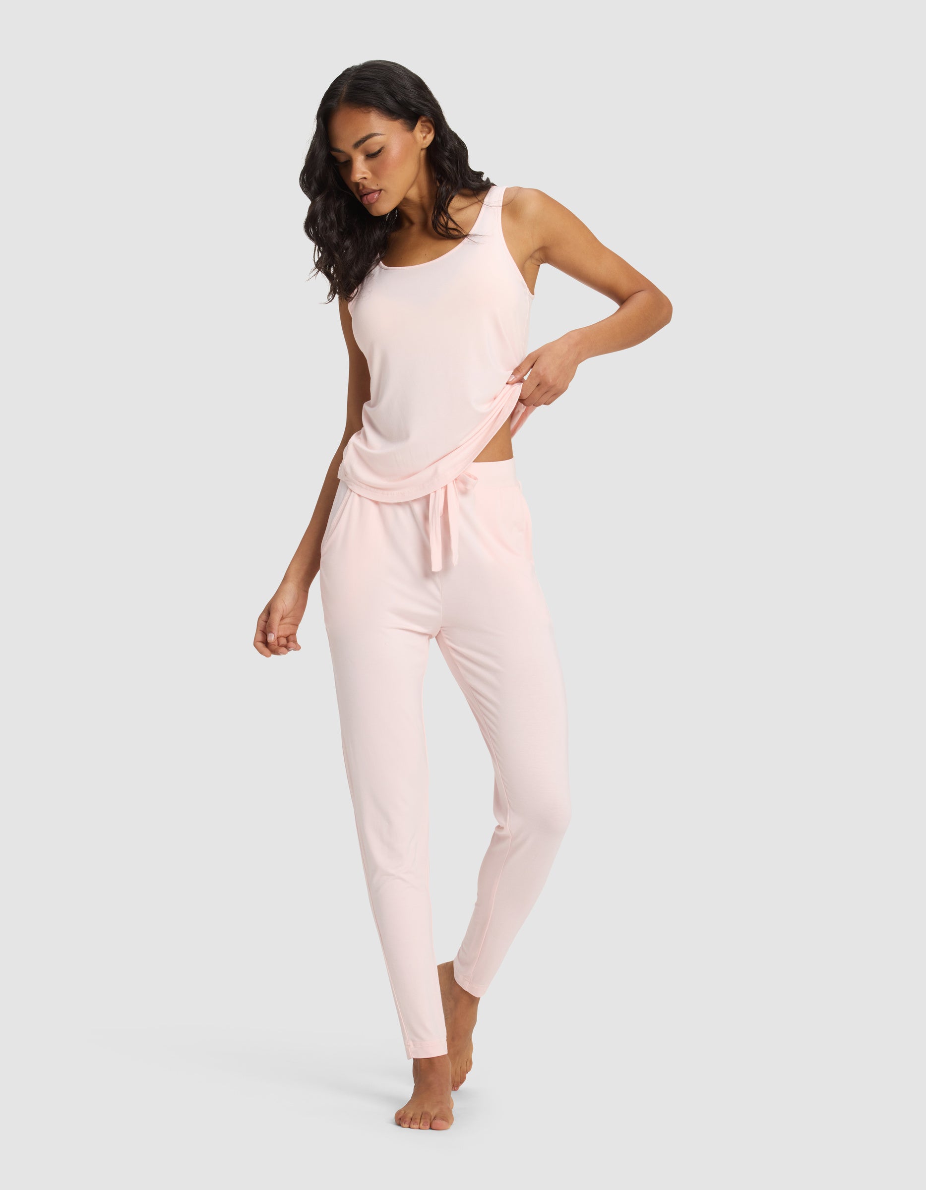 A woman models Cozy Earth's Women’s Bamboo Stretch-Knit Tapered Pant in light pink, barefoot on a plain background, lifting one side of her top and looking down with a relaxed expression.|Color:Blossom