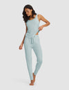A woman stands barefoot against a plain background, wearing the Cozy Earth Women's Bamboo Stretch-Knit Tank & Tapered Pant Set in light blue, looking down and gently lifting the hem of her top with one hand.|Color:Brook