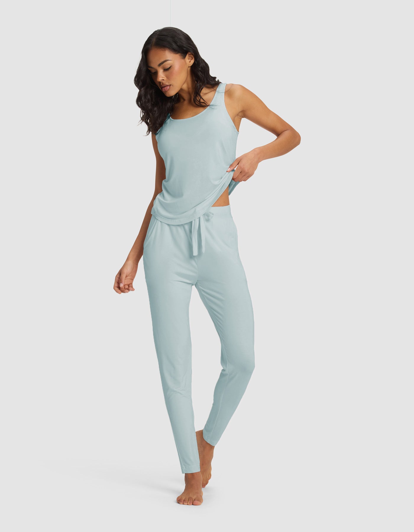 A woman stands barefoot against a plain background, wearing the Cozy Earth Women's Bamboo Stretch-Knit Tank & Tapered Pant Set in light blue, looking down and gently lifting the hem of her top with one hand.