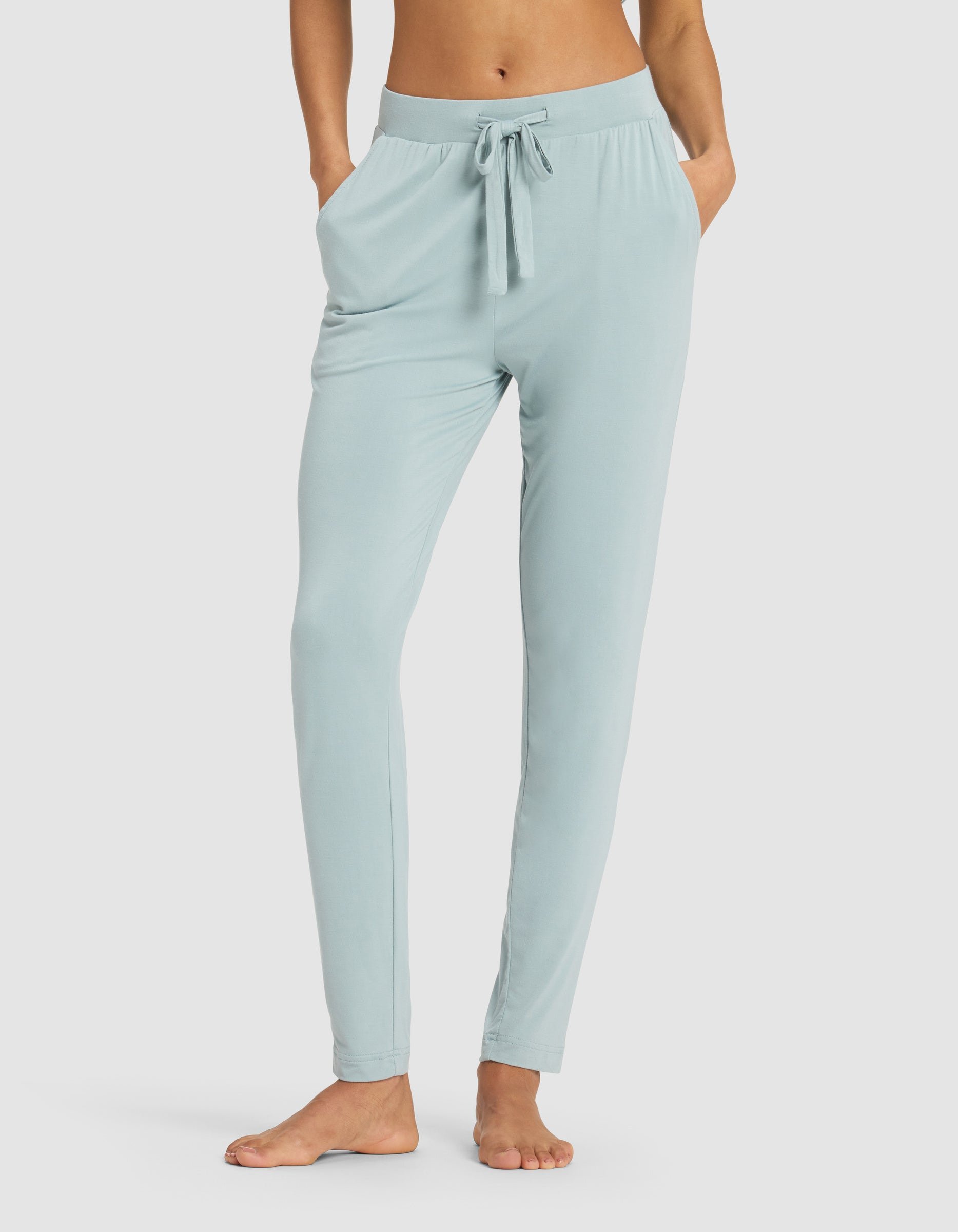 A woman models the Cozy Earth Women's Bamboo Stretch-Knit Tapered Pant in light blue, standing barefoot with hands in pockets against a plain white background.|Color:Brook