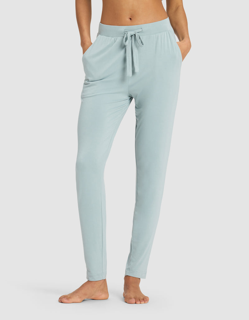 A woman models the Cozy Earth Women's Bamboo Stretch-Knit Tapered Pant in light blue, standing barefoot with hands in pockets against a plain white background.