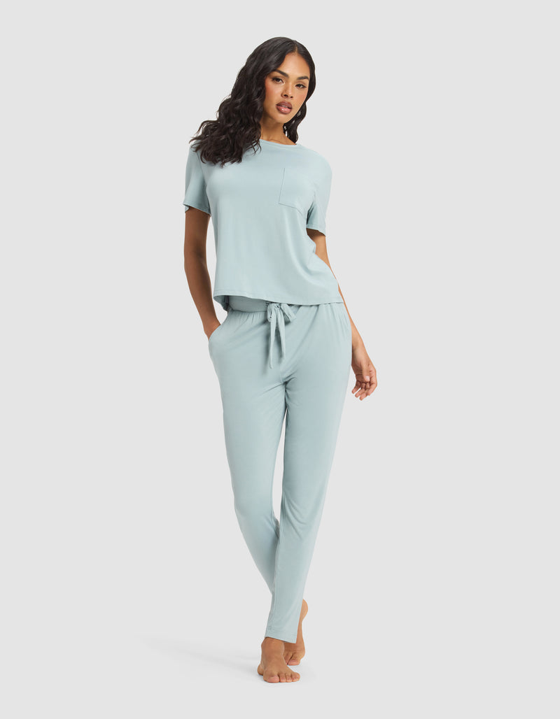 A woman with long dark hair poses barefoot on a white background, wearing Cozy Earth's Women's Bamboo Stretch-Knit Tapered Pant in light blue, paired with a matching short-sleeve top. One hand is in her pocket.