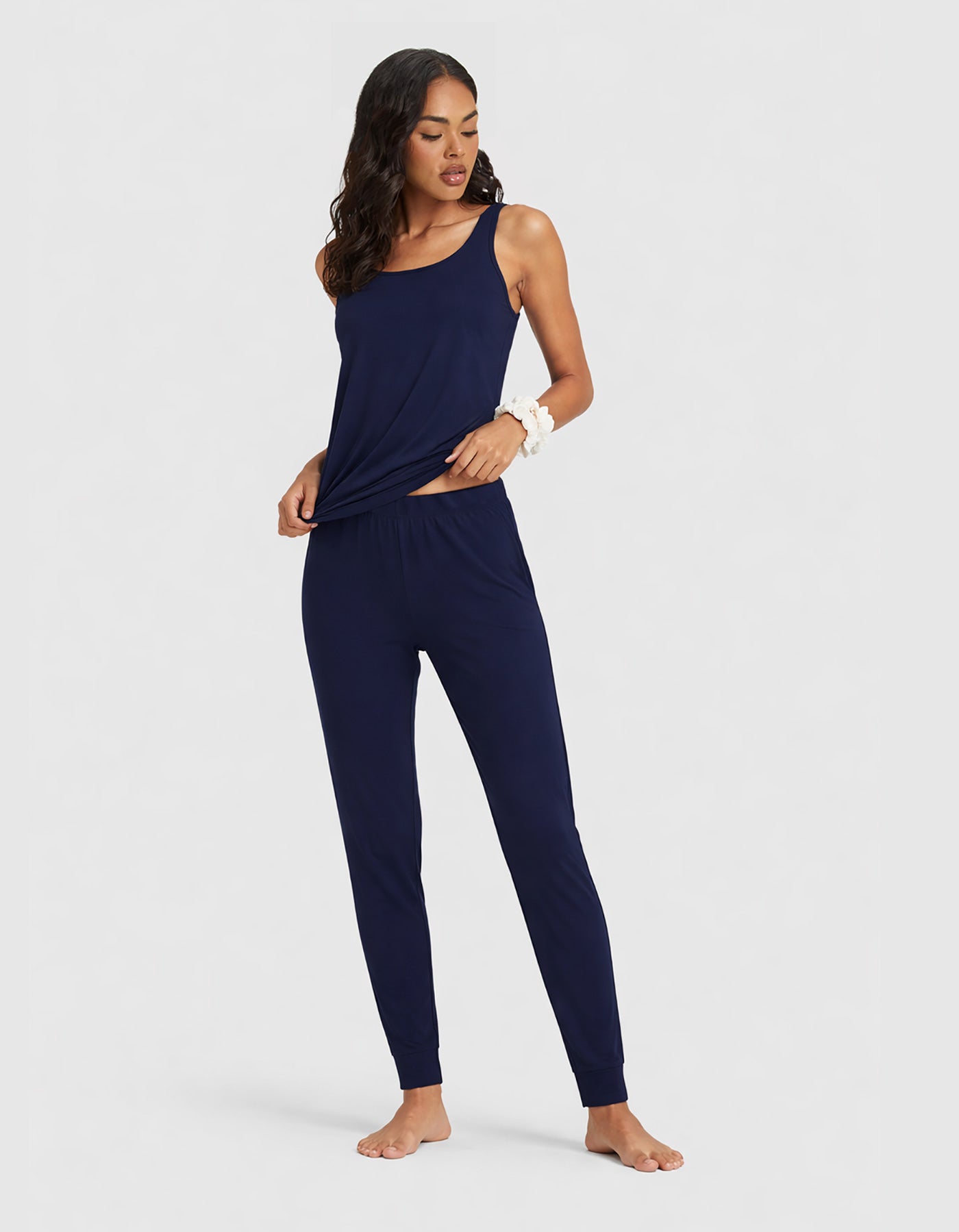 A woman poses barefoot in the Cozy Earth Women's Bamboo Stretch-Knit Tank & Tapered Pant Set, featuring a navy blue sleeveless top and matching jogger pants, against a plain light gray background.