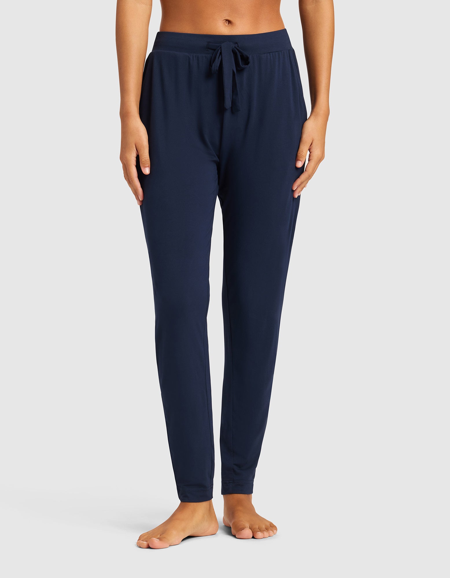 A woman models Cozy Earth's Women's Bamboo Stretch-Knit Tapered Pant in navy, shown barefoot with only her lower body visible against a plain light grey background.