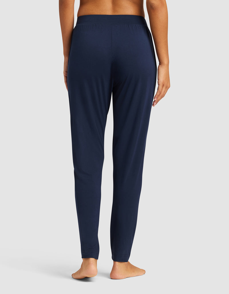 A barefoot woman, shown from the back, wears Cozy Earth's Women's Bamboo Stretch-Knit Tapered Pant in navy blue—a full-length, fitted pajama with an elastic waistband—against a plain light background.